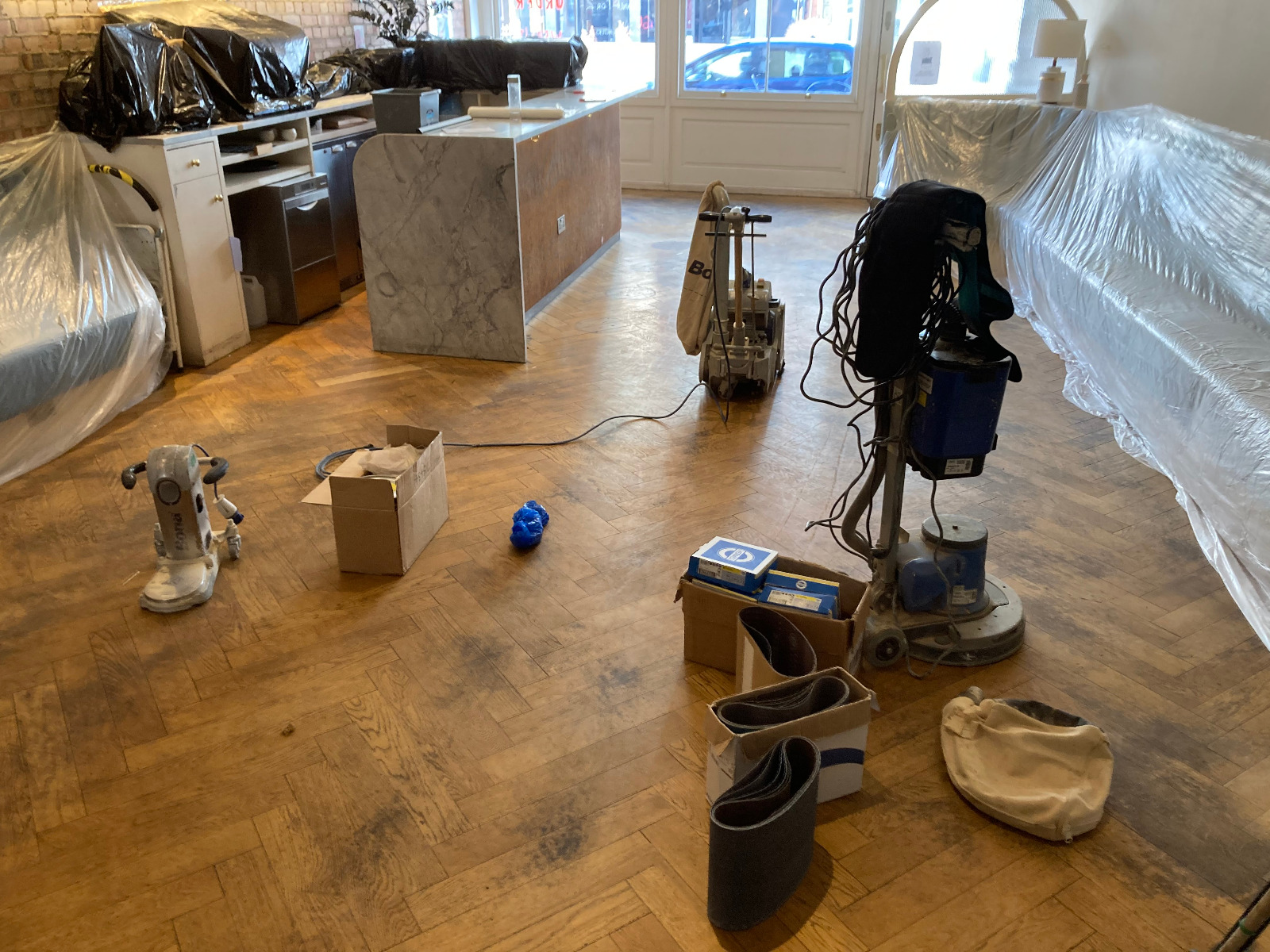 Sanding Parquet Floors in Sydenham – pre-sanding setup with belt and rotary machines and dust sheets around a worn herringbone oak parquet floor.