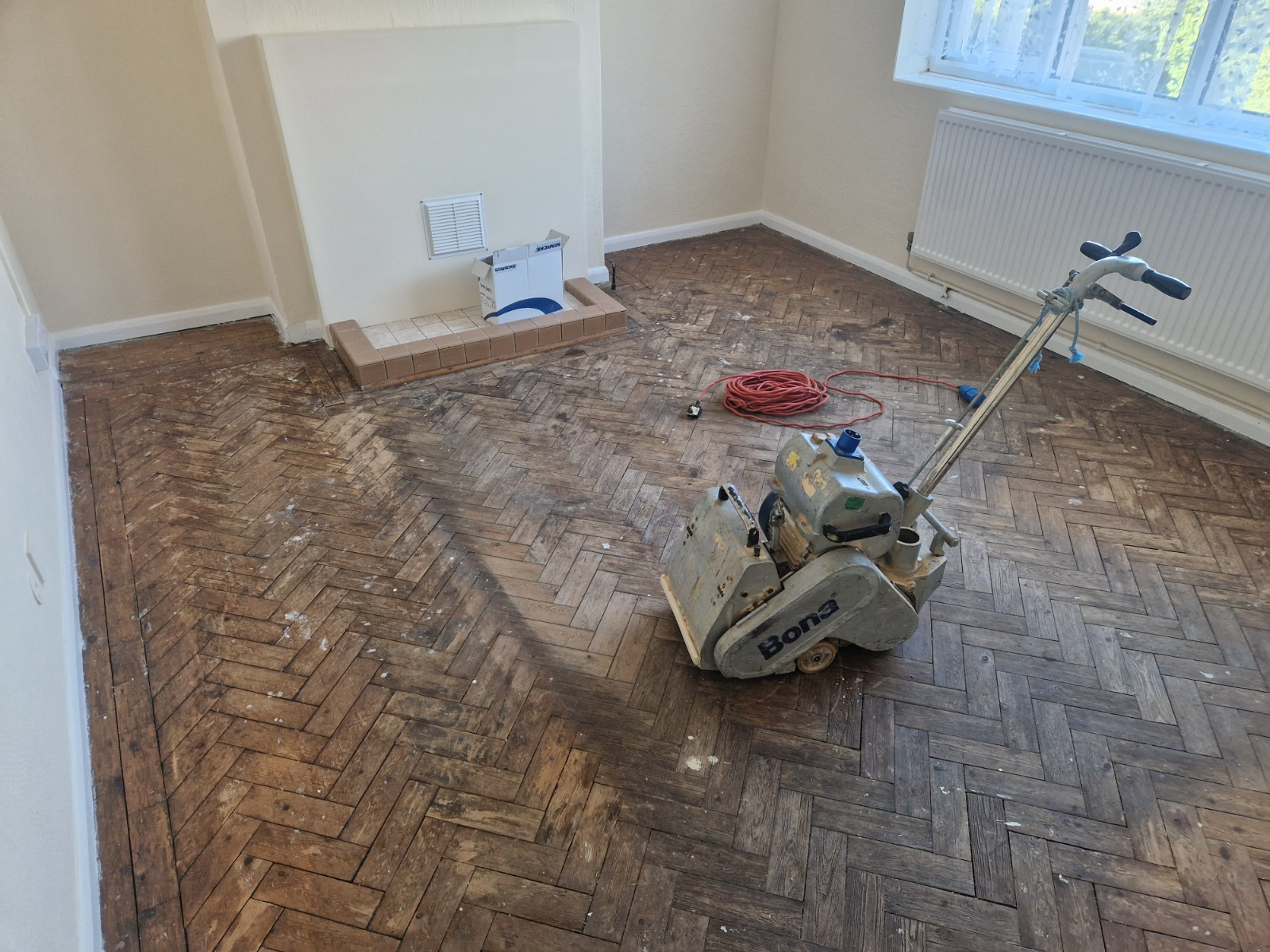 Wood Floor Sanding in Sydenham on an oak herringbone parquet living room floor, with a Bona belt sander starting to restore the worn, dark blocks.