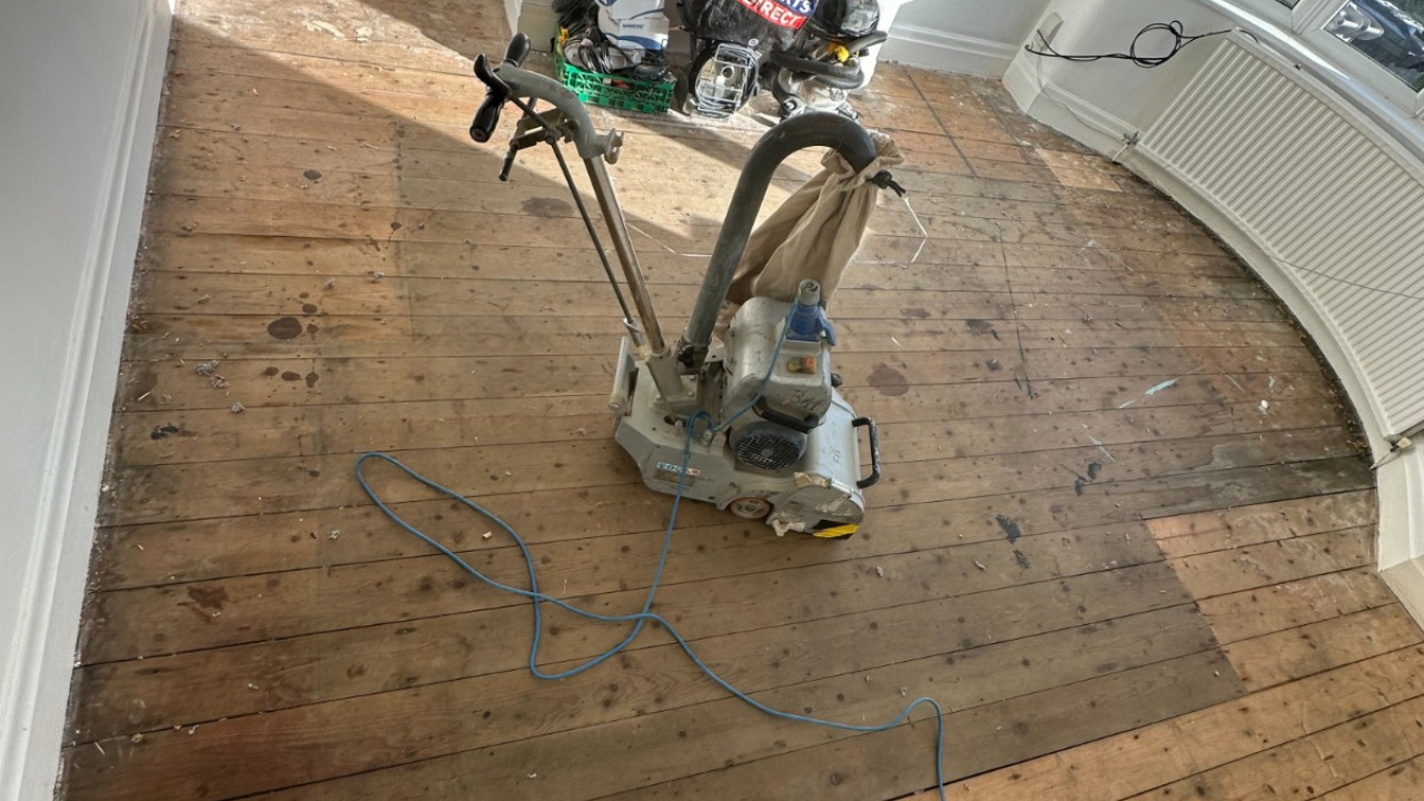 Wood Floor Sanding in Sydenham on original pine floorboards in a bay-window living room, with a professional drum sander preparing the worn boards for refinishing.