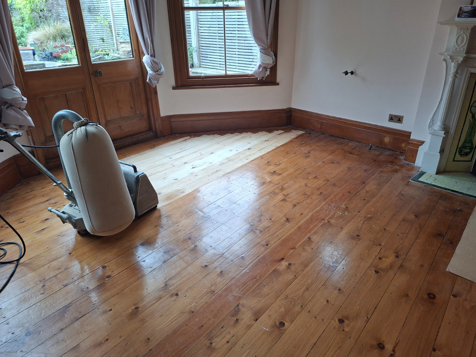 Wood Floor Sanding in Sydenham on softwood pine floorboards in a living room, with a drum sander revealing a lighter, freshly sanded section near the doors.